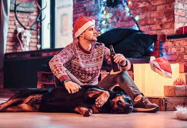 Un joven elegante sosteniendo una botella de cerveza mientras se sienta con su lindo perro en una sala de estar decorada en Navidad.