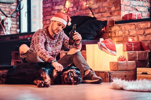 Un joven elegante sosteniendo una botella de cerveza mientras se sienta con su lindo perro en una sala de estar decorada en Navidad.