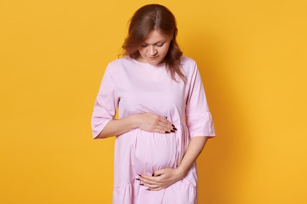 joven embarazada aislada en amarillo, adorable dama de pie con las manos sobre su abdomen, mira hacia abajo sobre su vientre, imagina a su futuro bebé. Concepto de maternidad