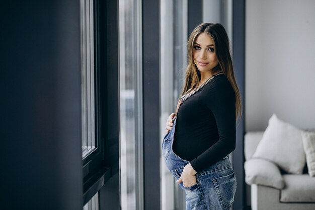 Joven embarazada sosteniendo su vientre y de pie junto a la ventana