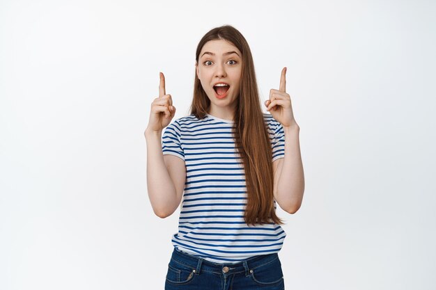 Una joven emocionada grita de asombro, señala con el dedo hacia arriba, muestra un anuncio de venta, se para sobre fondo blanco