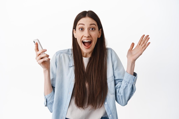 Una joven emocionada que sostiene un teléfono inteligente jadeando sorprendida de ganar en línea recibe noticias maravillosas en el teléfono de pie feliz contra el fondo blanco
