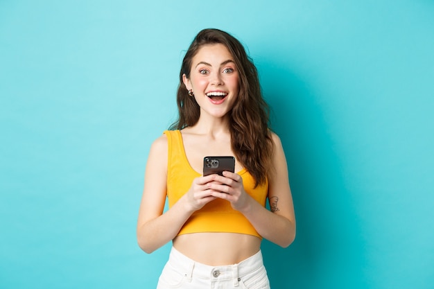 Joven emocionada recibe buenas noticias en el teléfono, sosteniendo el teléfono inteligente, mirando asombrado a la cámara con una sonrisa alegre, de pie contra el fondo azul.