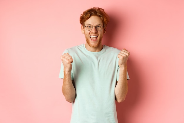 Joven emocionado ganando el premio, gritando de alegría y triunfo, haciendo puñetazos y diciendo que sí, de pie sobre un fondo rosa.