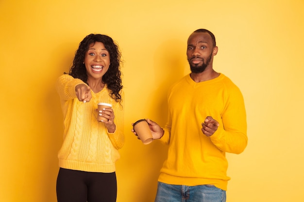 Joven emocional afroamericano y mujer en ropa casual brillante sobre fondo amarillo. Hermosa pareja. Concepto de emociones humanas, expresión facial, relaciones, anuncio. Beber café y señalar.