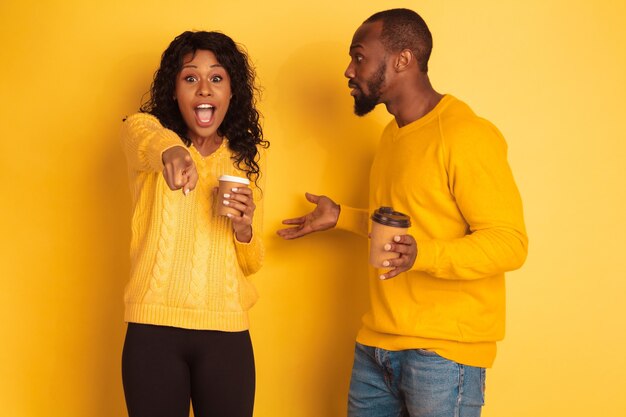 Joven emocional afroamericano y mujer en ropa casual brillante sobre fondo amarillo. Hermosa pareja. Concepto de emociones humanas, expresión facial, relaciones, anuncio. Beber café y señalar.