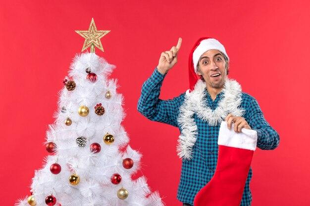 Joven emocional con sombrero de santa claus en una camisa azul a rayas y sosteniendo la Navidad apuntando arriba