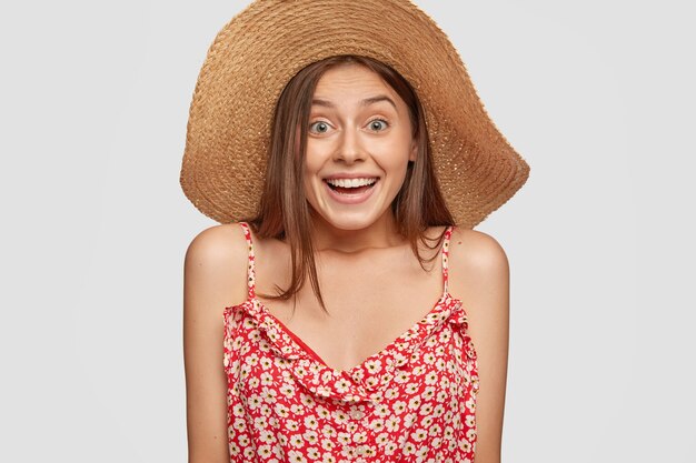 La joven emocional tiene una expresión alegre, usa un sombrero de paja de verano y un vestido de moda