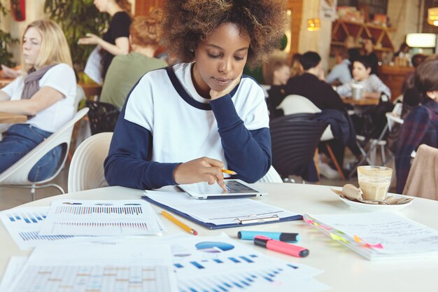 Joven empresaria africana con expresión seria y concentrada sentada en un café de coworking con un panel táctil y papeles, analizando información financiera en la tableta, apoyando el codo sobre la mesa