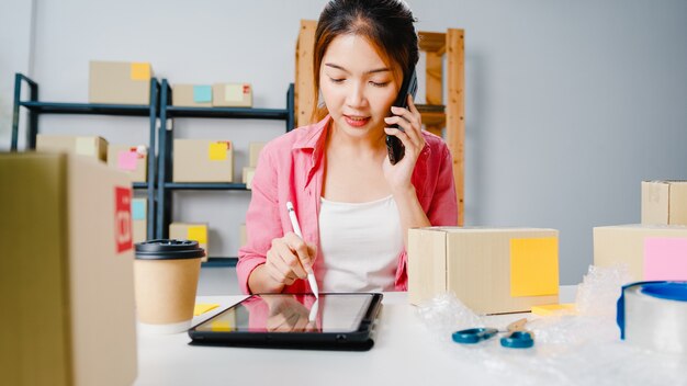 Joven empresaria de Asia mediante llamada de teléfono móvil recibiendo orden de compra y verificar el producto en stock, trabajar en la oficina en casa Propietario de una pequeña empresa, entrega de mercado en línea, concepto independiente de estilo de vida.