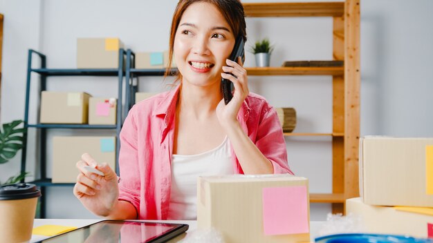 Joven empresaria de Asia mediante llamada de teléfono móvil recibiendo orden de compra y verificar el producto en stock, trabajar en la oficina en casa Propietario de una pequeña empresa, entrega de mercado en línea, concepto independiente de estilo de vida.