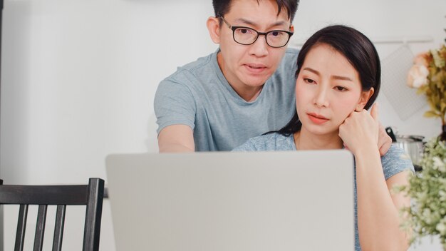 Joven empresaria asiática grave, estrés, cansado y enfermo mientras trabajaba en la computadora portátil en casa. El marido la consuela mientras trabaja duro en la cocina moderna de la casa por la mañana.