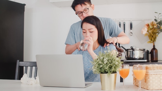 Joven empresaria asiática grave, estrés, cansado y enfermo mientras trabajaba en la computadora portátil en casa. El marido le da un vaso de agua mientras trabaja duro en la moderna cocina de la casa por la mañana.