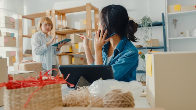 Joven empresaria asiática con mascarilla usando teléfono móvil recibiendo orden de compra y comprobando el producto en el trabajo de stock en la oficina en casa