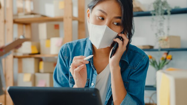 Joven empresaria asiática con mascarilla usando teléfono móvil recibiendo orden de compra y comprobando el producto en el trabajo de stock en la oficina en casa