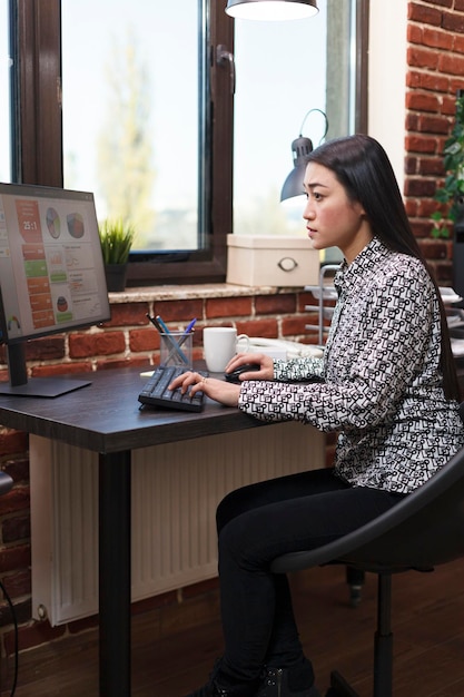 Joven empresaria asiática productiva revisando el gráfico de gestión de proyectos de negocios mientras está en la oficina de la empresa de marketing. Trabajador de la agencia financiera que revisa los resultados de la auditoría de inicio mientras se encuentra en un espacio de trabajo moderno.