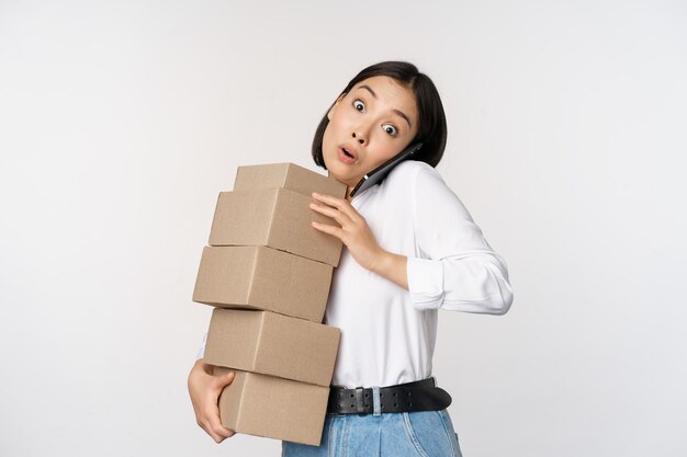 Una joven empresaria asiática responde una llamada telefónica hablando por teléfono móvil mientras lleva un montón de cajas con pedidos sobre fondo blanco.