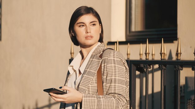 Joven empresaria atractiva con teléfono inteligente que parece pensativa en la calle de la ciudad. Chica elegante de pie al aire libre