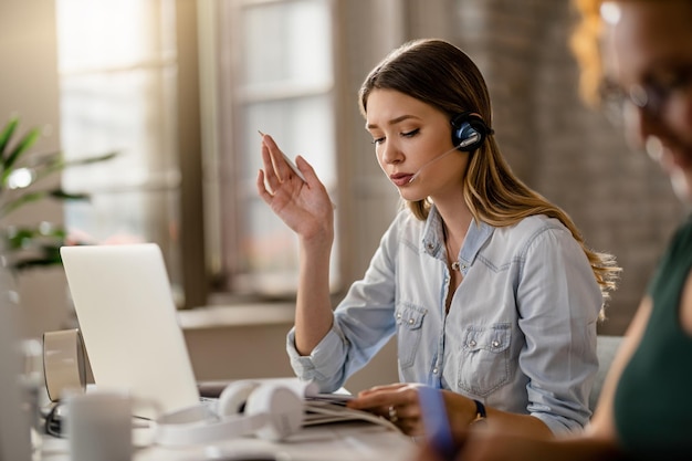 Joven empresaria con auriculares y comunicándose con un cliente mientras revisa el papeleo en la oficina