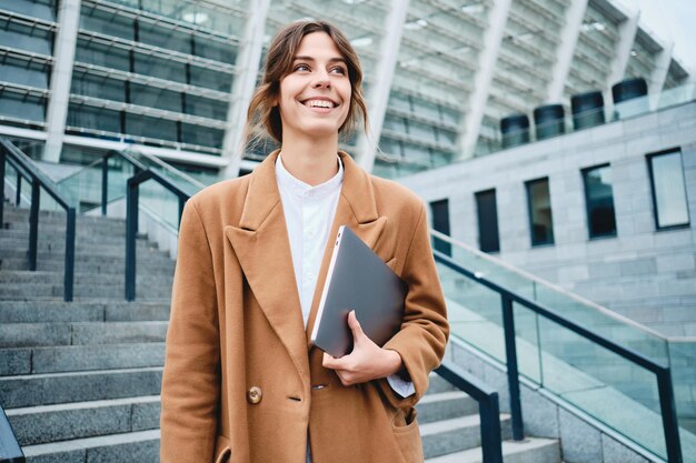 Joven empresaria bastante positiva en abrigo con portátil mirando alegremente al aire libre