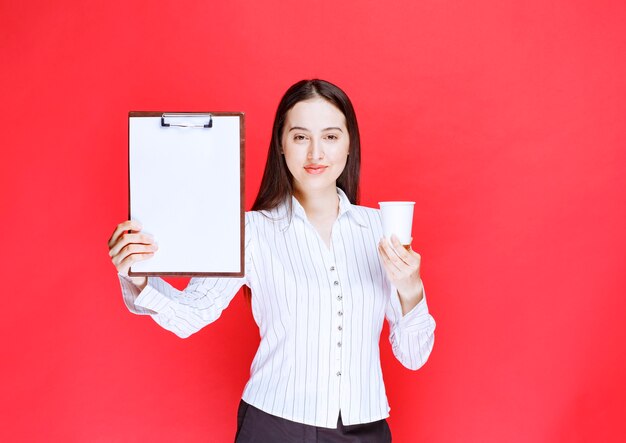 Joven empresaria bonita posando con portapapeles vacío y vaso de plástico.