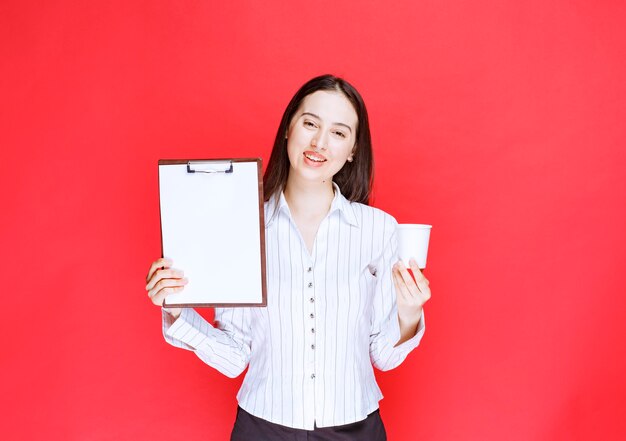 Joven empresaria bonita posando con portapapeles vacío y vaso de plástico.