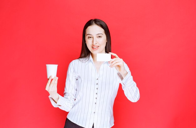 Joven empresaria bonita sosteniendo un vaso de plástico y una tarjeta de visita sobre fondo rojo.