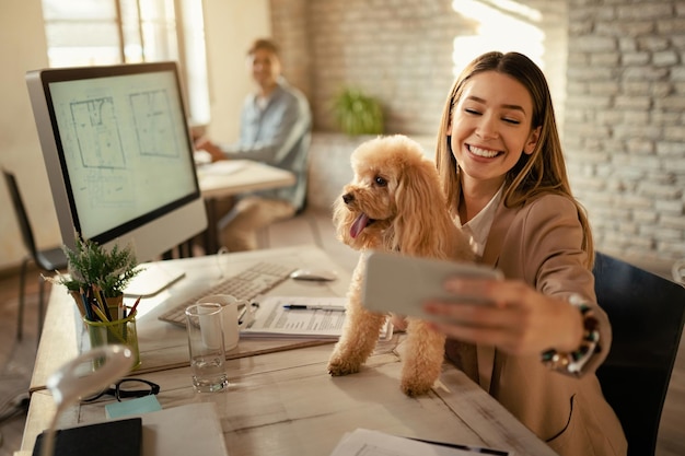 Joven empresaria divirtiéndose con su caniche mientras usa el teléfono y se toma selfie en la oficina