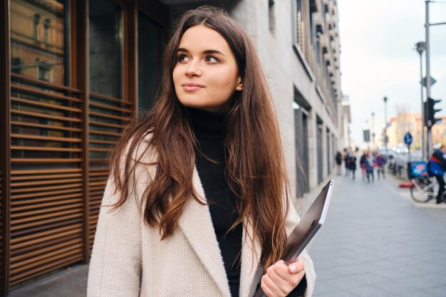 Joven empresaria con estilo con portátil mirando con nostalgia a la calle de la ciudad