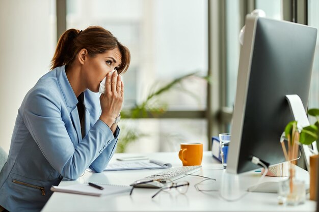 Joven empresaria estornudando en un pañuelo mientras trabajaba en el escritorio de su oficina