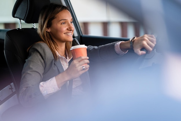 Joven empresaria feliz con café para ir conduciendo un coche