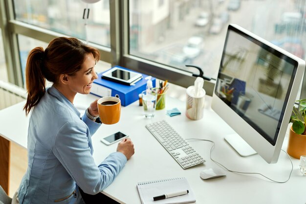 Joven empresaria feliz trabajando en una computadora y tomando café en la oficina