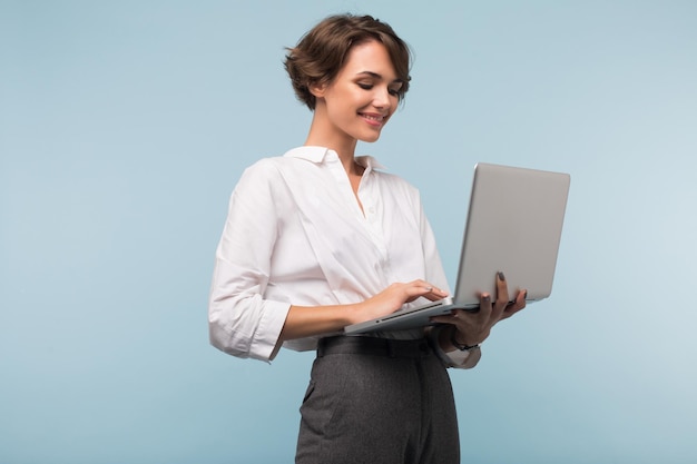 Joven empresaria hermosa con cabello corto oscuro en camisa blanca trabajando felizmente en una laptop sobre fondo azul aislado