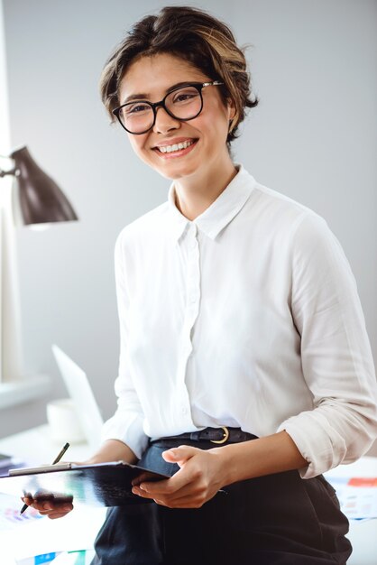 Joven empresaria hermosa celebración de carpeta, sonriendo en el lugar de trabajo en la oficina.