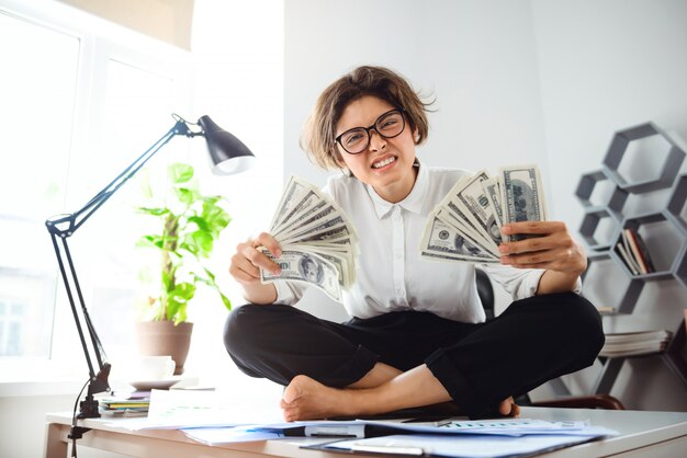 Joven empresaria hermosa con dinero, sentado en la mesa en el lugar de trabajo.