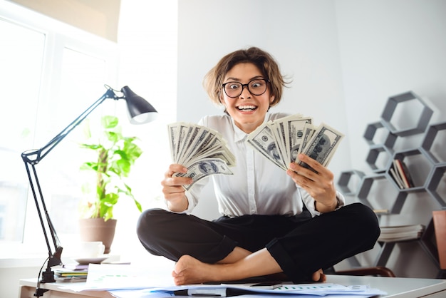 Foto gratuita joven empresaria hermosa con dinero, sentado en la mesa en el lugar de trabajo.