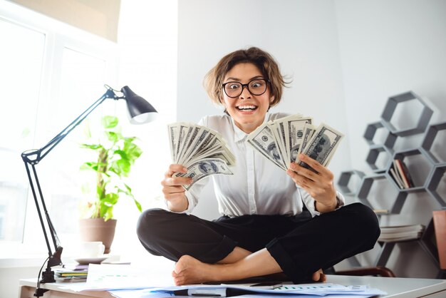 Joven empresaria hermosa con dinero, sentado en la mesa en el lugar de trabajo.