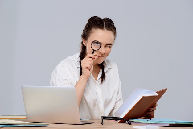 Joven empresaria hermosa sentada en el lugar de trabajo, con libro y lupa