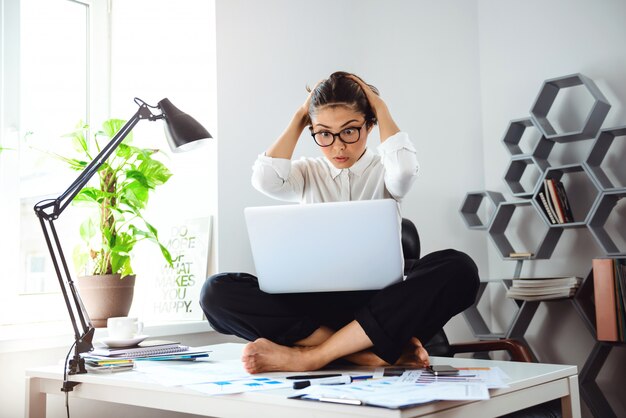 Joven empresaria hermosa sentada en la mesa con el portátil en el lugar de trabajo.