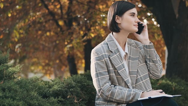 Joven empresaria hermosa vestida con un blazer a cuadros hablando por teléfono inteligente durante el trabajo en la calle de la ciudad Tecnología moderna