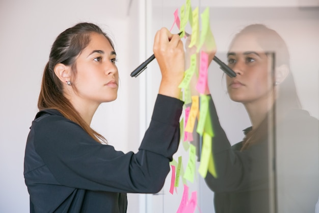 Joven empresaria latina escribiendo en etiqueta con marcador. Centrado confiado hermosa morena mujer gerente compartiendo idea para proyecto y tomando nota. Concepto de lluvia de ideas, negocios y formación