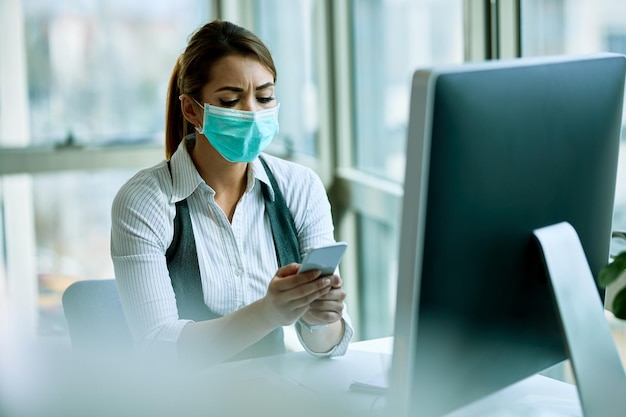 Joven empresaria con máscara facial enviando mensajes de texto en un teléfono inteligente mientras trabaja en la oficina