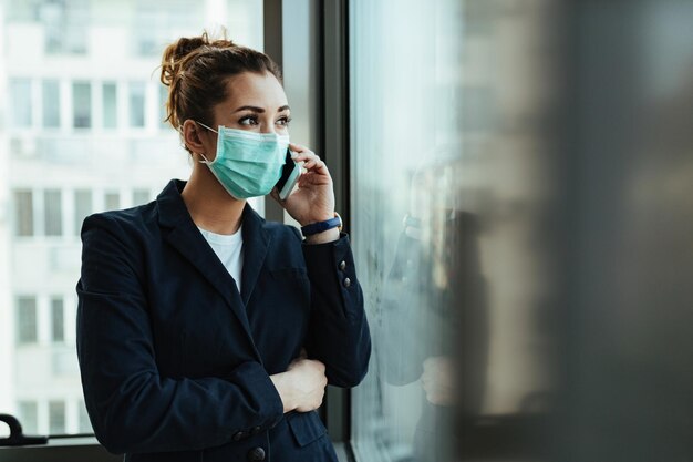 Joven empresaria con mascarilla parada junto a la ventana y hablando por teléfono