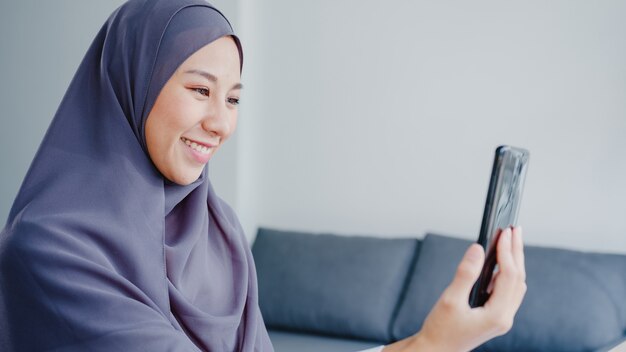 Joven empresaria musulmana de Asia que usa un teléfono inteligente, habla con un amigo por videochat y hace una lluvia de ideas en línea mientras trabaja de forma remota desde casa en la sala de estar.