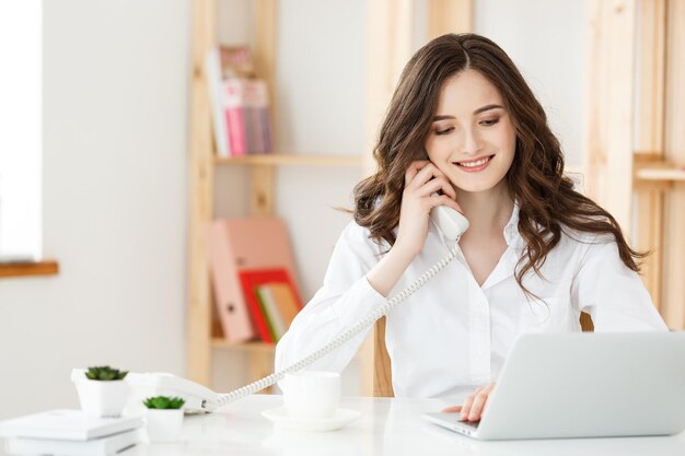 Joven empresaria o secretaria sentada en el escritorio y trabajando Sonriendo y mirando a la cámara