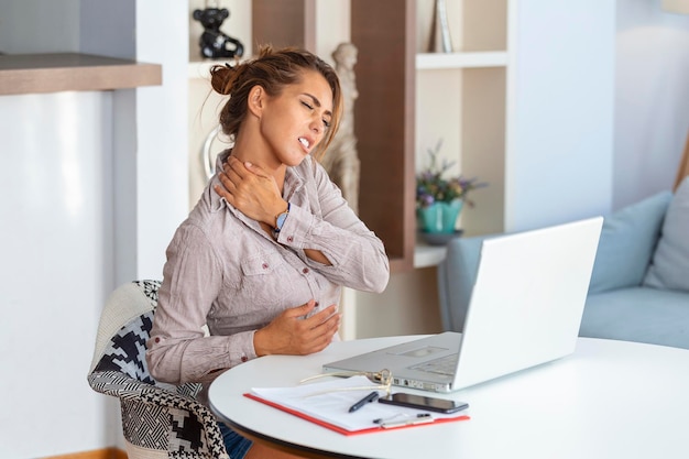 Joven empresaria que sufre de dolor de cuello masajeando su cuello mientras está sentada en su lugar de trabajo en la oficina del hogar