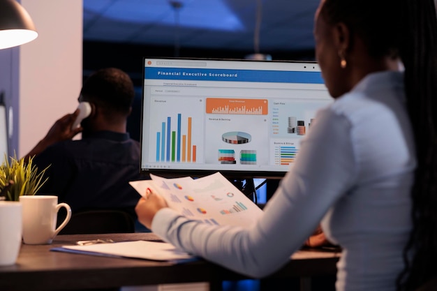 Joven empresaria que trabaja con gráficos estadísticos en el monitor de la computadora, analizando los datos de la empresa sobre el papeleo. Uso de información en línea para crear un informe de investigación antes de la fecha límite, por la noche fuera del horario de atención.