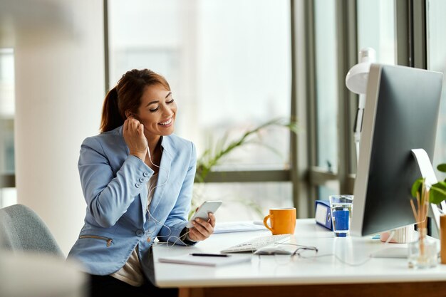 Joven empresaria relajándose mientras usa el teléfono móvil y escucha música con auriculares en la oficina