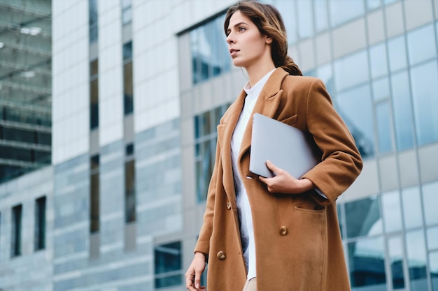 Joven empresaria seria y elegante con abrigo y portátil caminando pensativamente por las calles de la ciudad