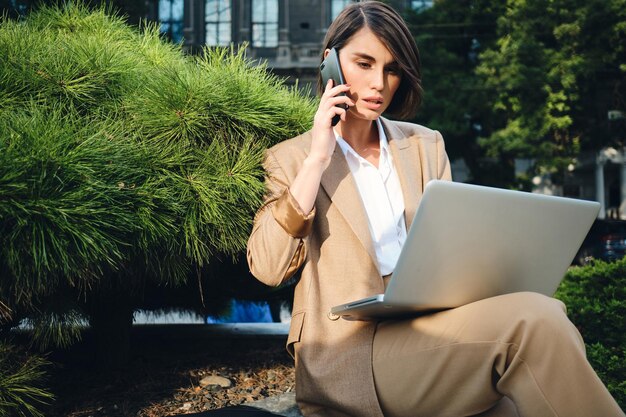 Joven empresaria seria y elegante hablando por teléfono celular y trabajando en una computadora portátil al aire libre